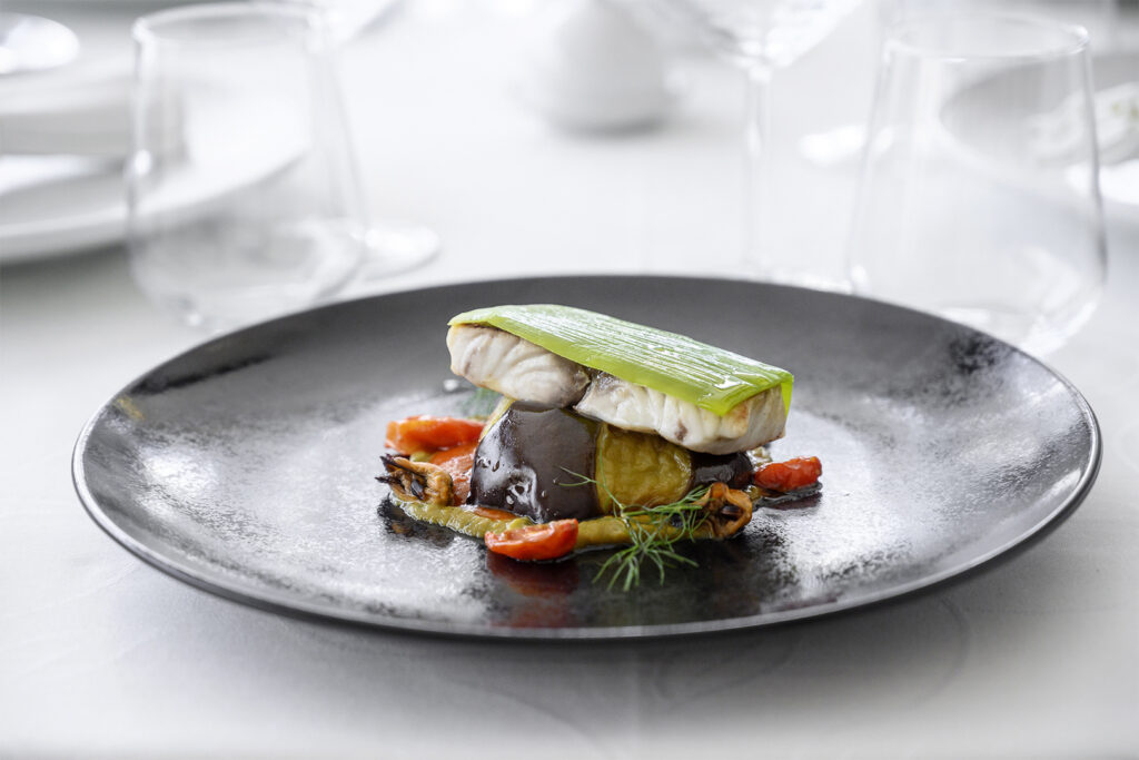 Elegante piatto gourmet con filetto di pesce, verdure grigliate e porro glassato, servito su un piatto nero in un ristorante raffinato in Sicilia