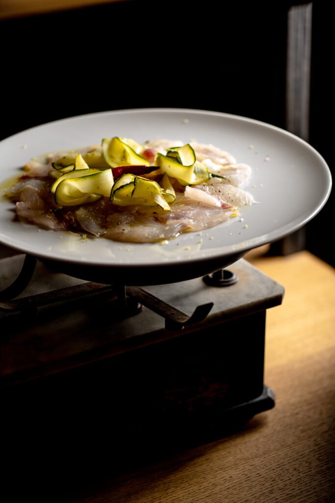 Carpaccio di pesce con zucchine su piatto bianco, esempio di foto di un piatto firma per il sito web di un ristorante siciliano, foto di Andrea Ventura